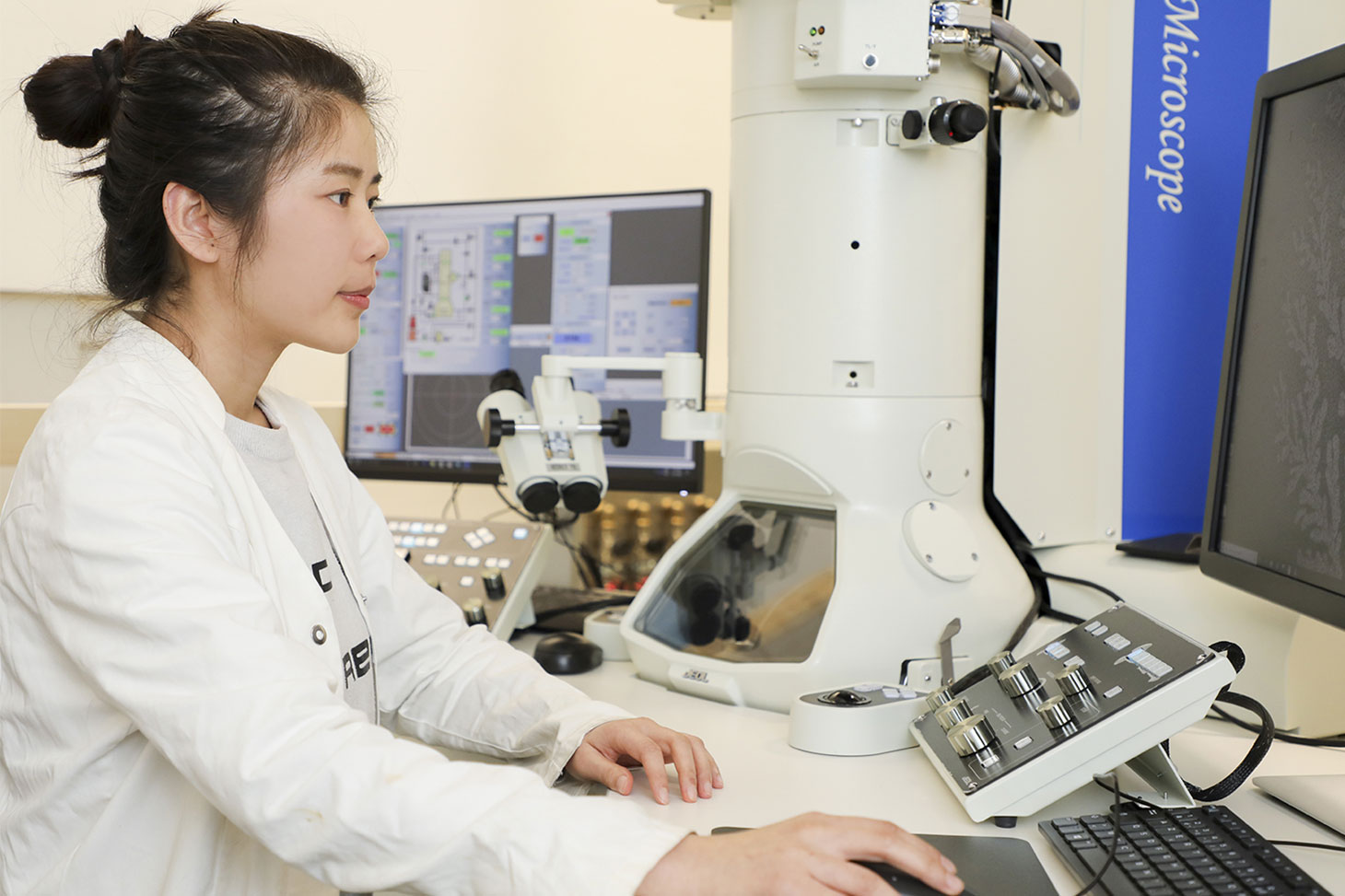 Une jeune chercheuse en blouse blanche travaille devant un ordinateur et un microscope dans un laboratoire